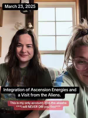 The image is a selfie-style video still featuring two young women. The woman on the left is the predominant focal point, with long, dark hair and a slight smile, set against a backdrop of a home interior with wooden details. The woman on the right is partially visible, wearing glasses and a high bun. Text overlays include a date and a message about ascension energies, and a visit from aliens.