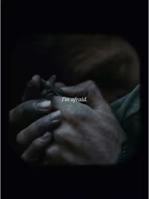 A close-up shot focuses on a pair of hands, dark in tone, holding a small object. The hands are the primary focus, with the background obscured in shadow. The lighting is dim, creating high-contrast shadows across the hands' surface and the object.