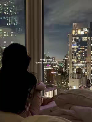 A nighttime view from a high-rise apartment window. The primary focus is a silhouette of a person sitting on a bed, gazing out at a city skyline filled with illuminated buildings. Text is overlaid on the image.
