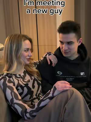 A woman with long blonde hair is sitting on a couch with a man. The man has short dark hair and is wearing a black hoodie. The woman is looking to her left and the man is looking at the woman on his right and they seem to be cozy.