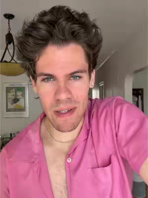 A selfie taken from the chest up features a man with dark, tousled hair. He's wearing a pink, open-collared shirt. The background includes glimpses of a home interior.
