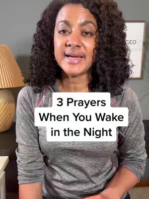 A woman with curly dark hair stands facing the camera, speaking. A white rectangular overlay with black text, "3 Prayers When You Wake in the Night," covers her midsection. She is wearing a gray and pink long-sleeved shirt and is positioned in front of a dark brown couch and a lamp to her left.