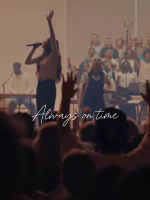 A live performance with a female singer center stage, arms raised, singing into a microphone, with a large chorus on the right and a crowd with raised hands in the foreground. Soft lighting and a warm color palette evoke a sense of worship and community.