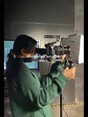 A person in a green sweater holds a large camera with accessories in in a dark area. Text overlays show the phrase “If I could get out of this place”.