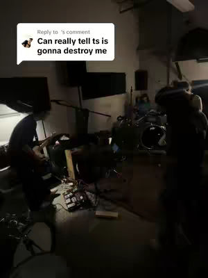 The image shows a dimly lit room, likely a music studio or practice space. The main focus is on a drummer in the center, facing the drums. Various equipment, including amplifiers, mixers, and cables, is scattered around the space, adding to the impression of a working environment. The lighting is low, and there are a few people in the background.