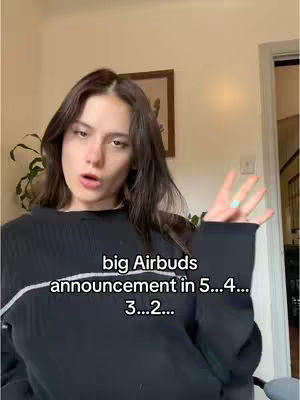 A young woman with long dark hair is centered in the frame, looking at the camera with a slightly open mouth and raised eyebrows. She is wearing a black sweater. Text overlayed on the image counts down 'big Airbuds announcement in 5...4...3...2...'. The background suggests an indoor setting, with a beige wall visible, behind and to the side of the woman, there is greenery and a door frame slightly visible on the right.