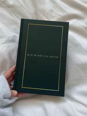The image shows a closed, dark green book on a white bedspread. The book has a gold border and the text 'READ ME WHEN YOU NEED ME' in a lighter color. A hand, wearing a gray sleeve, holds the book from the left.