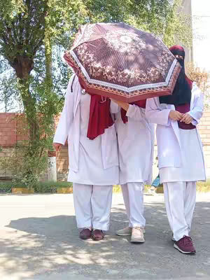 Three people, potentially medical students, are standing on a paved area, partially obscured by a large, patterned umbrella. The individual on the right wears a hijab. They're all dressed in white medical attire, and the scene appears to be outdoors with hints of greenery in the background.