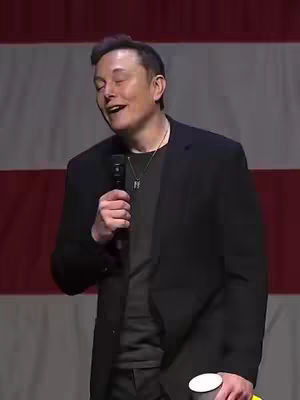Elon Musk is standing on a stage, holding a microphone and a cup, with a backdrop featuring a red and white striped design similar to an American flag. He is smiling slightly with his eyes closed, and he is wearing a black jacket, a gray t-shirt, and a silver necklace.