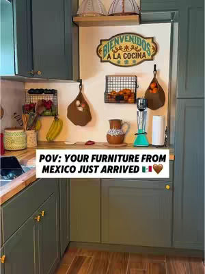 A kitchen corner with green cabinets and wooden countertops. A sign that says Bienvenidos A La Cocina hangs above a countertop with kitchen tools and fruit baskets.