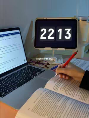 The image shows a laptop open on a desk alongside an open book, a digital clock displaying 22:13, glasses, and a hand holding a pen as if writing. The scene is lit with warm, soft lighting, suggesting a study environment. The composition is typical of a study setup, with the laptop screen visible, likely displaying study material or notes.