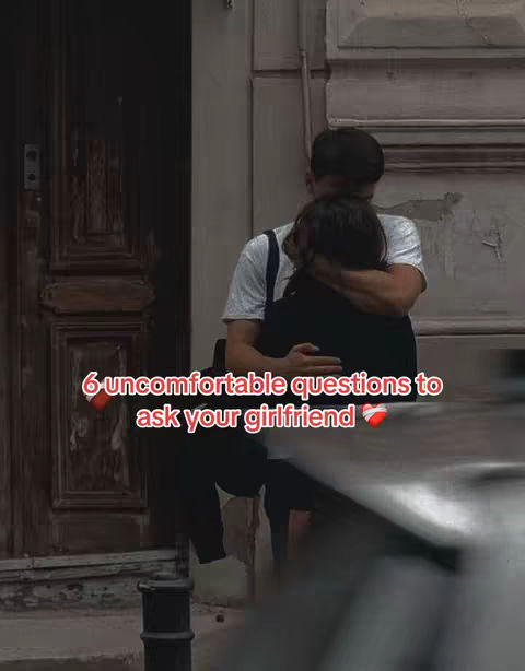 A man and woman are embracing outdoors. The woman appears to be leaning against him, and the background is blurred, with a building behind them. Text overlayed in red states, '6 uncomfortable questions to ask your girlfriend' with hearts at the end.