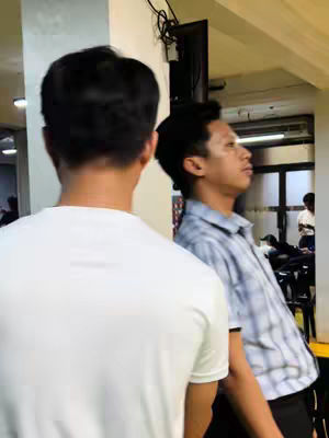 Two young men stand indoors, likely at a religious gathering or church. The slightly blurred background suggests the photo was captured during an event. The image is composed with the back of one person in the foreground while another person is standing next to him.