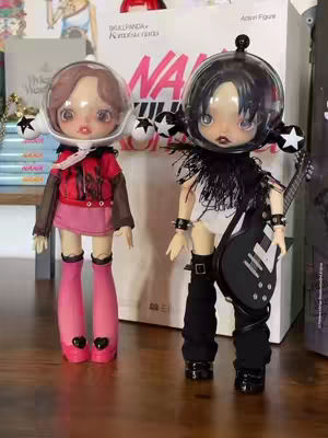 Two collectible dolls are displayed on a wooden surface. The doll on the left is dressed in a red jacket and pink skirt, with a white headpiece and transparent face covering. The right doll is wearing a black and white outfit with a guitar. Behind them is a box with the word 'NANA' on it.
