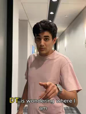 A man in a pink t-shirt is pointing at the viewer, standing in a hallway with a neutral background.