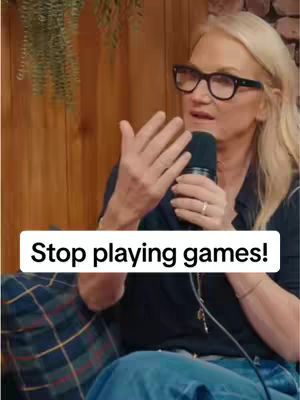A woman with blonde hair and glasses is holding a microphone and speaking. She is wearing a dark shirt and blue jeans, and there's a white rectangle overlay with the text "Stop playing games!". In the background there is a wooden wall and what appears to be a plant.