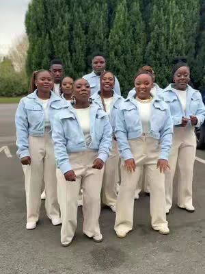 A group of people, primarily women, are standing in a row, posing for a photo. They appear to be dressed in a matching uniform, with light blue jackets and light-colored pants. The background consists of lush green trees. They are facing the camera, and it appears like there is a slight smiling expression.