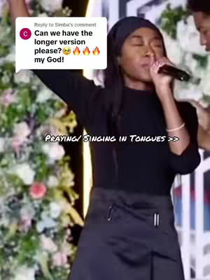 A woman with long, dark hair is singing into a microphone, wearing a black top and skirt, set against a backdrop of white and green floral arrangements.