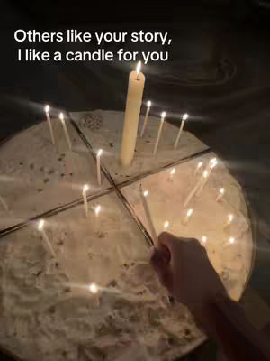 A dimly lit scene with a round surface, perhaps a table or a large plate, covered with what appears to be a textured cloth. A central, large candle burns brightly, and numerous smaller candles surround it, some lit, others not. A hand is reaching in to light one of the candles, with the overall effect creating a sense of ritual or reverence.