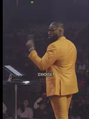The image shows a man in a yellow suit, standing on a stage, speaking. The text overlay at the bottom reads CHOISE.