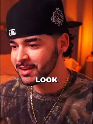 A close-up shot of a man with short dark hair, a trimmed beard, and a black baseball cap. The man is looking slightly downward with a soft expression. The word "LOOK" is superimposed on the image in bold white letters.