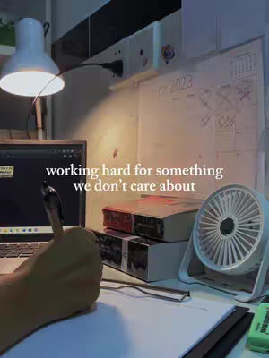 A desk setup with a laptop, books, a lamp, and a fan. Text overlayed says 'working hard for something we don't care about'. The scene is dimly lit, suggesting nighttime or a study environment.