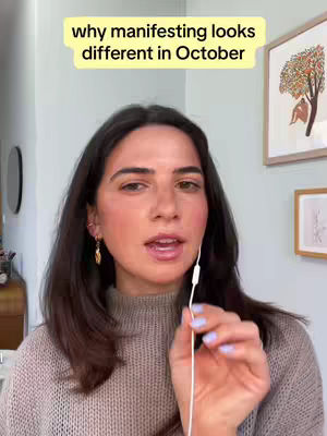 A woman with dark hair and fair skin is the central figure. She is wearing a gray sweater and appears to be indoors, possibly in a home setting. A text overlay reading, "why manifesting looks different in October" is displayed at the top. The background includes a neutral-colored wall, a round mirror, and framed artwork.