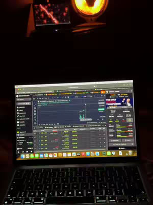 A dark room is lit by the glow of a computer screen, a small monitor, and a spotlight. The laptop is open, displaying a trading platform interface with charts and data. The background is blurred, suggesting a focused and dimly lit environment.