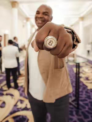 An out-of-focus shot of a man in a business setting. He is smiling, wearing a blazer, a white shirt, and dark pants. His fist is in the foreground, displaying a silver ring with a Bitcoin logo.