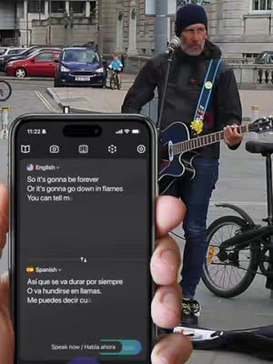 A man plays guitar and sings on a city street, the screen of a phone displaying text translation from English to Spanish is held in front of the scene.
