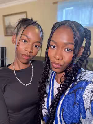 Two young women are positioned close together in a selfie. The woman on the left is wearing a black long-sleeve top and a silver necklace. The woman on the right is wearing a blue and white patterned sweater. Both have dark hair and are looking at the camera. The background shows a living room with neutral-toned walls and a framed picture. The lighting is soft and natural.