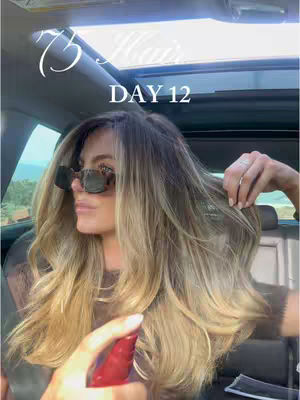 A woman with long blonde hair and sunglasses sits in a car. The sun is shining through the sunroof. She is holding out her hair and spraying it with hair product.