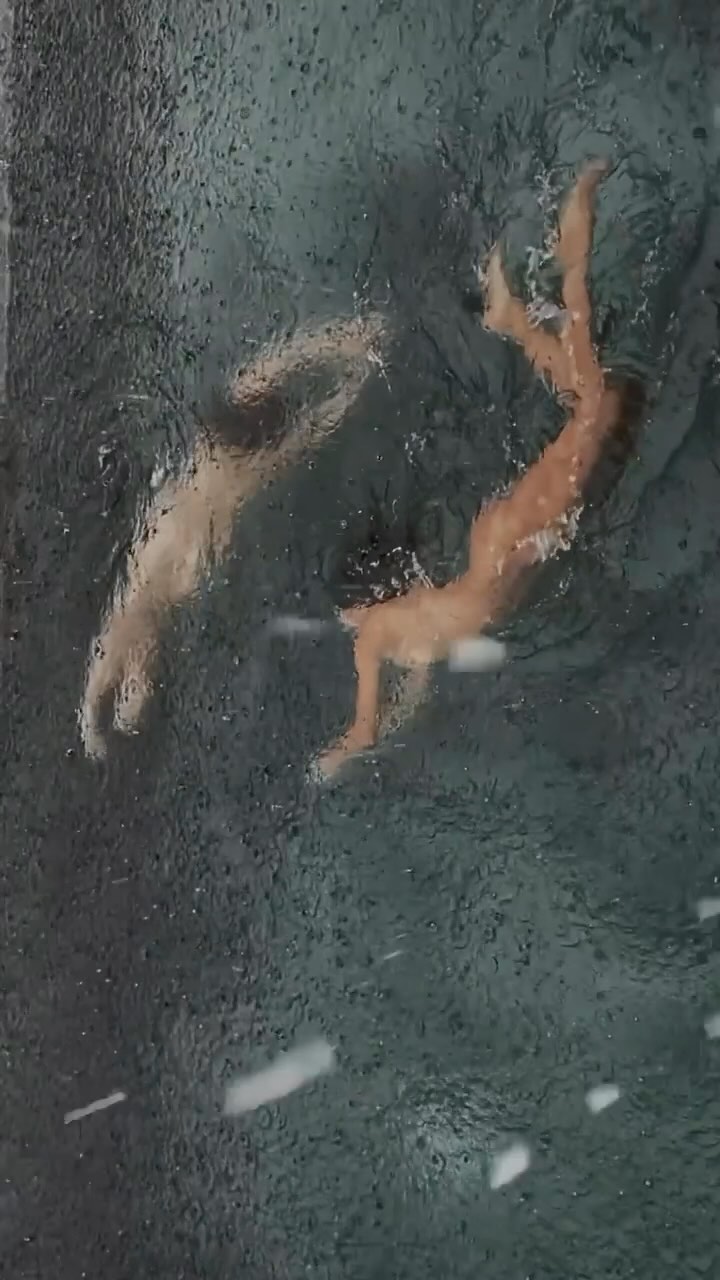 The cover image shows an overhead view of two people swimming in a body of water. The water is dark and textured, possibly due to rain or ripples. The swimmers are partially submerged, with their limbs visible.