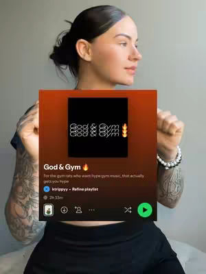 A woman with dark hair in a bun holds a Spotify playlist titled 'God & Gym'. She is facing slightly right, with tattoos visible on her arm. The playlist's cover is a dark orange gradient with text that says: God & Gym. The image has a casual, motivational feel.