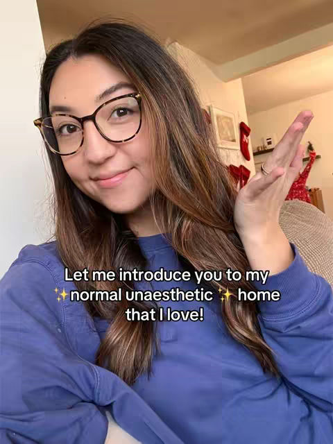 A selfie-style shot featuring a woman with long brown hair, glasses, and a blue sweater. She smiles warmly and gestures with her right hand. Text overlayed offers a welcoming message. The background is softly blurred, suggesting a home interior.