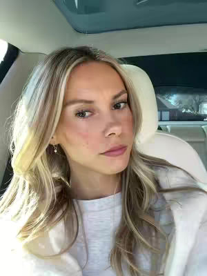 A woman with long blonde hair looks at the camera with a sad expression while sitting in a car. The background includes the car's interior and parts of the outside scenery.