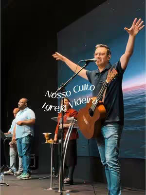 A man with glasses is on stage, holding a guitar and singing into a microphone. He has his arms raised. Behind him is a screen with a blurred image, and there are other people on the stage. In the foreground, there are some instruments and a glass stand.