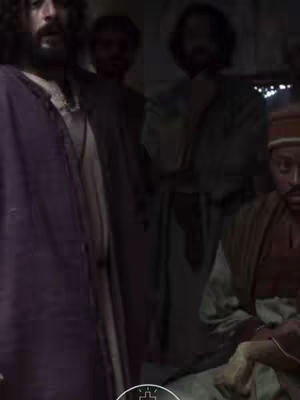 A scene from a film featuring multiple figures in a dimly lit interior. The central figure, dressed in a purple robe, appears to be the focus, with others partially visible in the background, illuminated with a low-key lighting scheme.