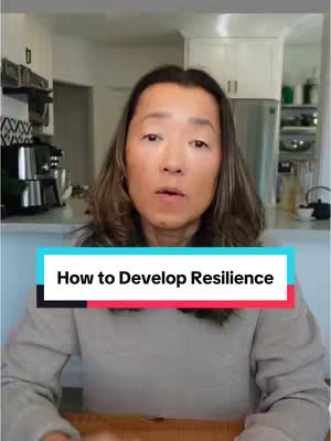 A woman with long, dark hair is centered in the frame against a blurred home interior backdrop. She is wearing a grey sweater and appears to be speaking directly to the viewer. A turquoise and white rectangular text overlay, reading 'How to Develop Resilience', is placed at the bottom-center of the frame.