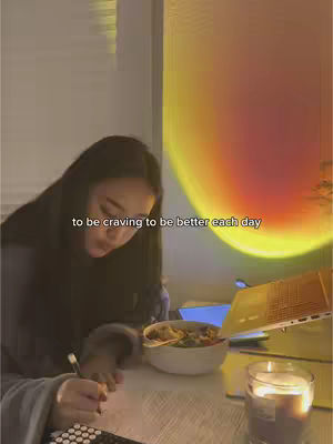 A cover photo showcases a young woman sitting at a desk, focused on writing. A bowl of food sits in front of her, and a candle and laptop stand on the desk as well. The main color profile is warm, emphasized by the glow from a sunset lamp in the background.