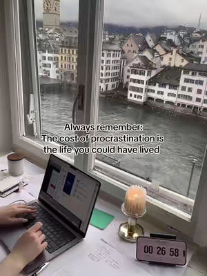 A person working on a laptop by a window overlooking a cityscape. A candle and a timer sits on the desk.