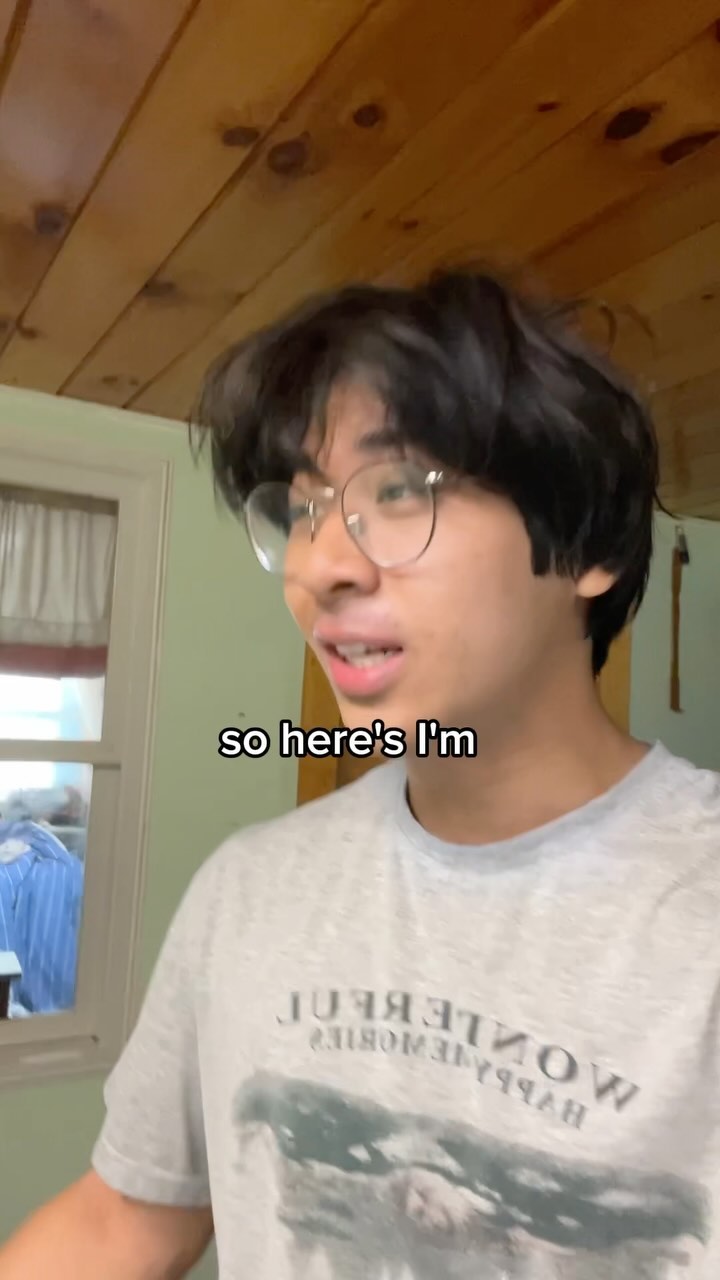 A young man with glasses and a grey t-shirt is shown from the chest up, positioned slightly to the left of the frame. A text overlay appears at the bottom, reading 'so here's'. The background features a wooden wall and a window. The lighting appears natural and bright.