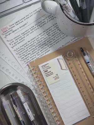The image captures a top-down view of a study setup. A blank checklist sticky note, a transparent ruler, and a ring-bound notepad are featured primarily. Documents with handwritten text in a serif font are also visible, along with pens in a metal holder. The color scheme is muted, with soft lighting.