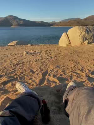 A first-person perspective shot from a sandy beach or cliffside overlooking a large body of water. The bottom part of the frame shows the feet of the person, wearing Converse shoes with the sun setting. In the middle there is a black bottle of beer, with legs sprawled from the middle of the shot to the bottom corners.