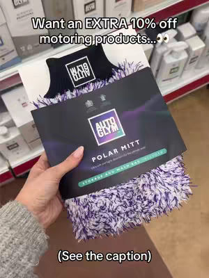 A woman's hand holds a black and purple car wash mitt on a shelf in a store. The mitt is in a black package with white text and a logo. Above the mitt is text in a black box that states: Want an EXTRA 10% off motoring products. The background shows shelves of various car cleaning products, some in white bottles. The overall tone is promotional.