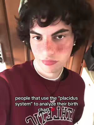 A young man with dark, curly hair and a burgundy sweatshirt is centered in a selfie-style shot. Freckles are visible on his lightly-colored skin. He wears a pearl necklace and has a neutral expression. The background is somewhat obscured, with hints of a wooden door or wall visible behind him. Text overlayed on the bottom: people that use the placidus system to analyze their birth chart.