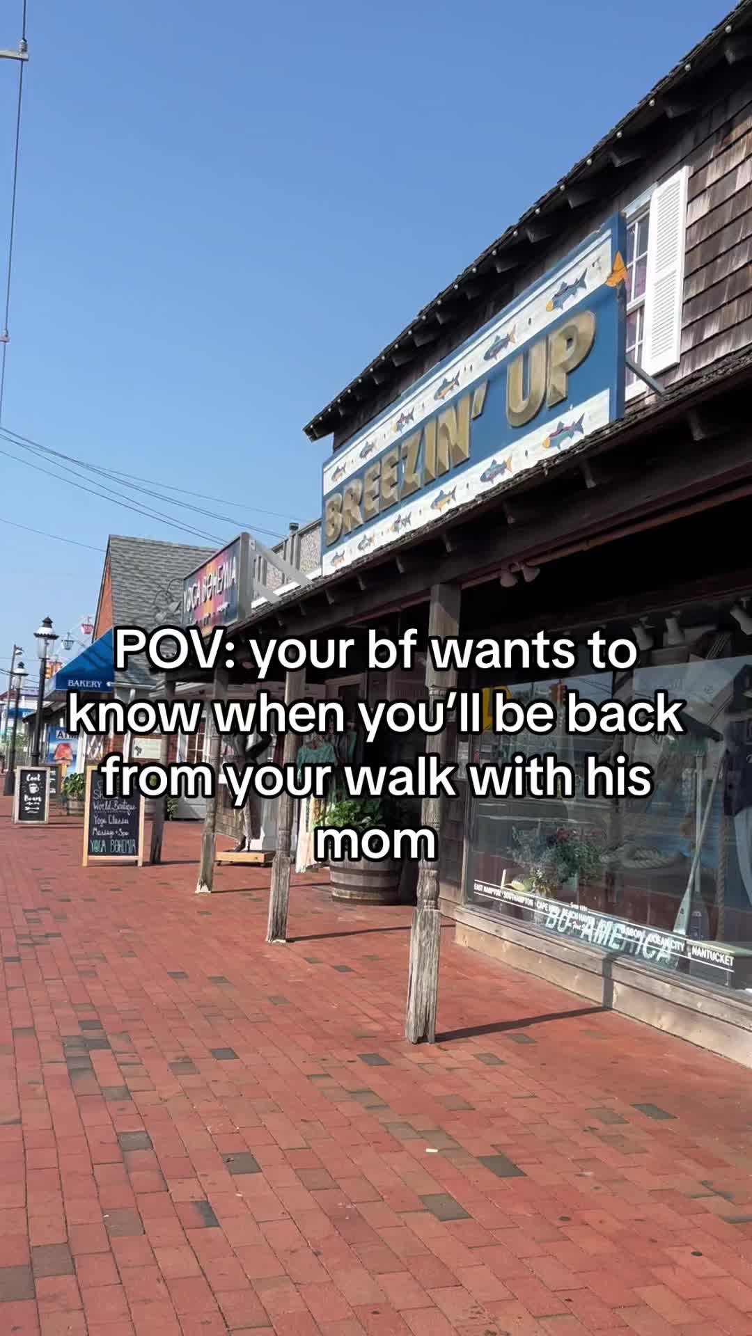 The cover image presents a vibrant outdoor scene, likely a street in a coastal town, on a sunny day. Red brick pavement fills the foreground, leading into the image with a pathway. On the left, there's a 'Bakery' sign, hinting at a local business. The buildings on the right feature a weathered exterior with wooden accents and a sign that says 'Breezin' Up.' A window display is visible at the bottom. Added text overlays appear, giving context while capturing the eye.