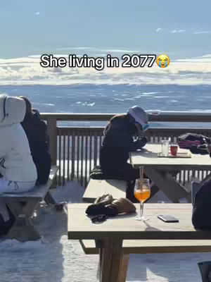 A person sits at a restaurant table outside wearing a jacket and headwear. The table has a drink and a cell phone on it and the background is a blurry landscape of snow and mountains.