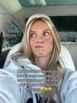 A woman is sitting in the driver's seat of a car, looking off to the side with a thoughtful expression. She has long blonde hair and is wearing a white hoodie. Text overlays on the image read "My boyfriend figured out how to send messages to my lockscreen whenever we're apart during long distance".