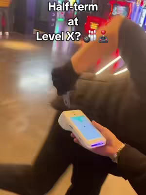 A person is holding a point-of-sale machine in front of an arcade or gaming venue. The person is wearing a dark jacket or sweater. The background is blurred, but one can see arcade games with bright lights.